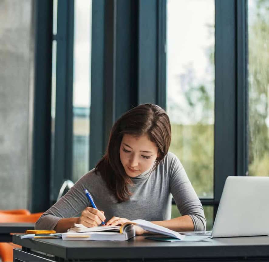 Young woman studying at a desk with a laptop and books in a bright room.